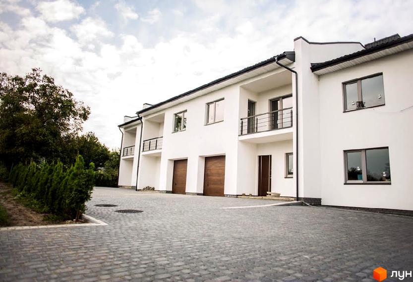 Зовнішній вигляд новобудови на вул. Щирецька, 117. Білі фасади з балконами та гаражними воротами. Прибудинкова територія з бруківкою та зеленими насадженнями.