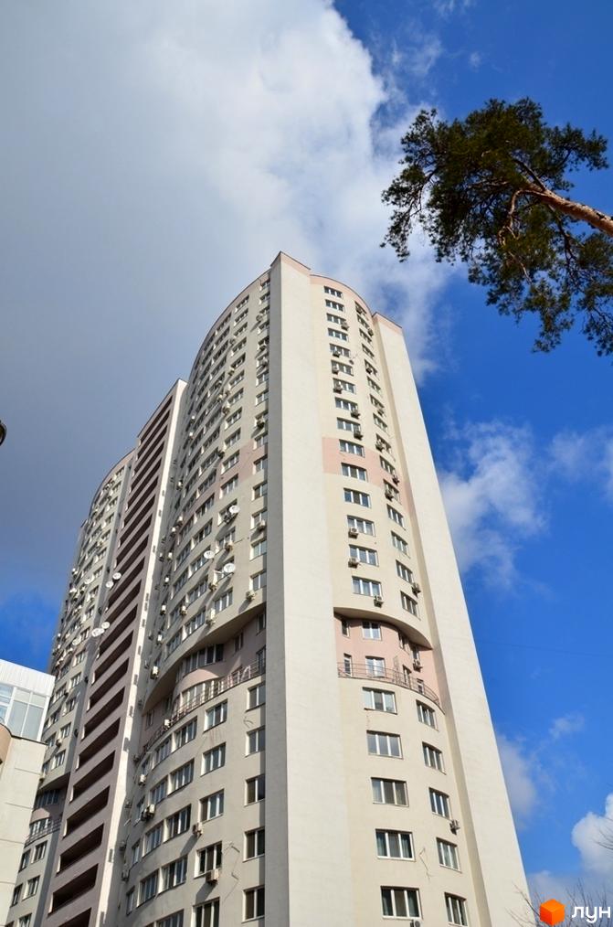 Житловий комплекс Вікторія: високий світлий будинок з вигнутим фасадом, численними вікнами, кондиціонерами та супутниковими антенами на тлі неба і дерева.