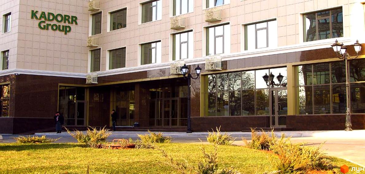 Фасад Житлового комплексу Перша Перлина виконаний у світлих тонах з великими вікнами та темною вхідною групою. Перед будинком розташований зелений газон з кущами.