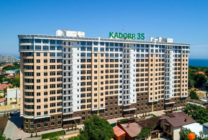 Зовнішній вигляд житлового комплексу Kadorr 35 з фасадом у бежевих та білих тонах. Навколо будівлі розташована зелена прибудинкова територія з деревами та невеликими будинками.