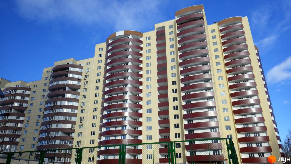 Багатоповерховий житловий комплекс Парковий з бежевим фасадом та округлими балконами коричневого кольору. На передньому плані видно зелену огорожу.