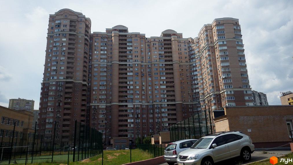 Багатоповерховий житловий комплекс Голосіїво з вигнутим фасадом світло-коричневого кольору та численними вікнами. На передньому плані видно парковку та спортивний майданчик.