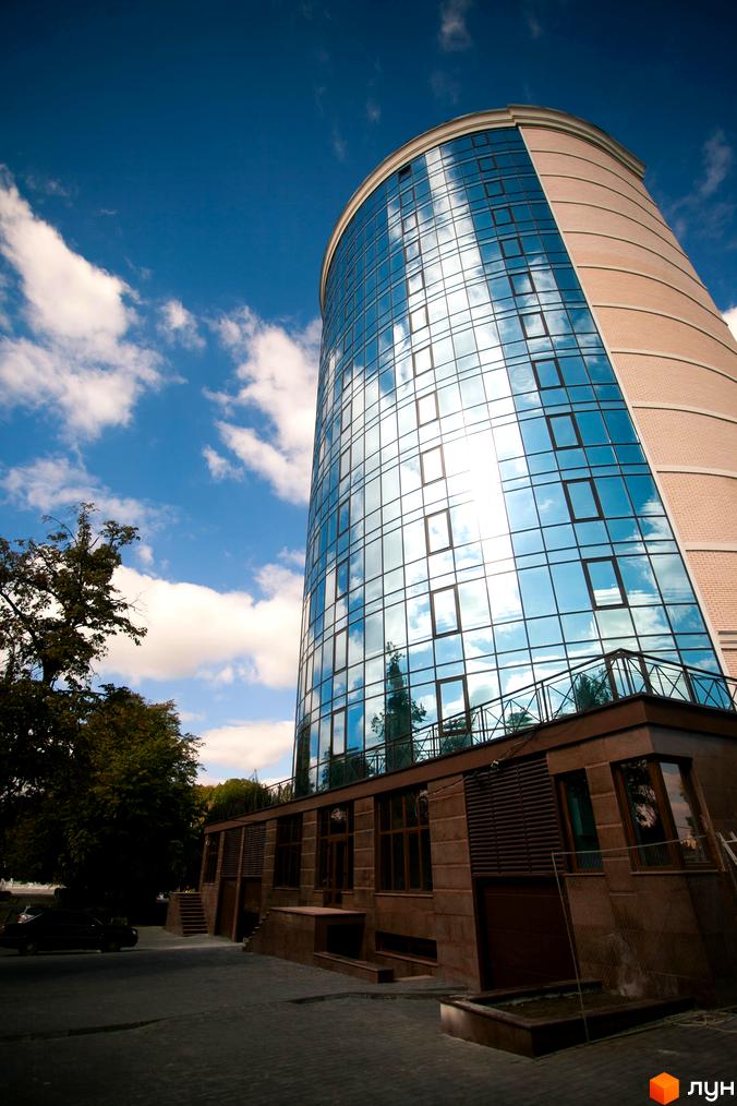 Житловий комплекс Paradise має вигнутий фасад з синього скла, що відбиває небо та хмари, і частину з світлої цегли. Навколо будівлі видно вимощену територію та дерева.