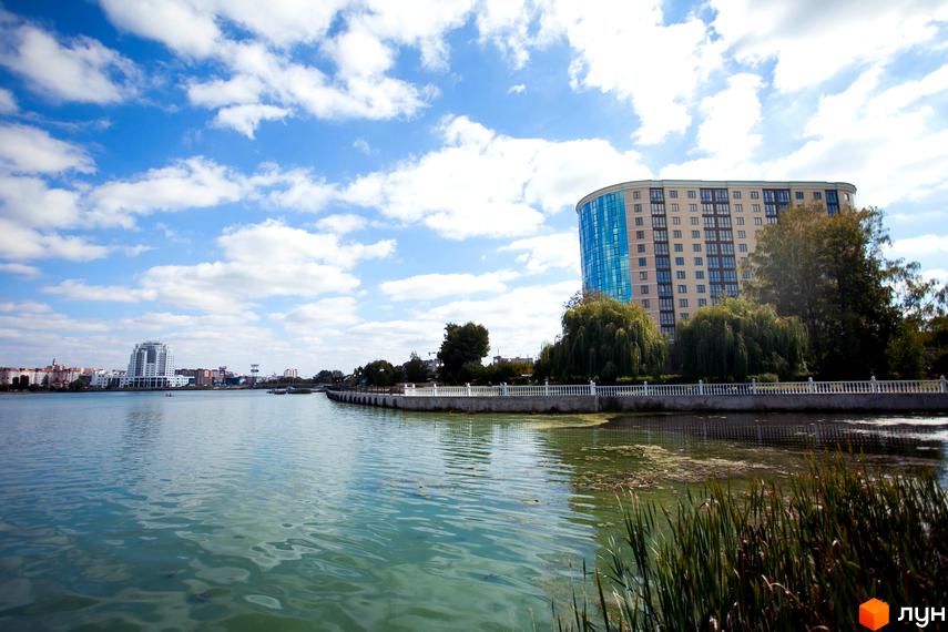 Зображення Житлового комплексу Paradise біля озера. Високий будинок з блакитними вікнами, зелені дерева та доглянута прибережна зона.