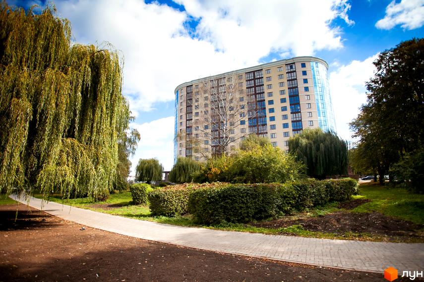 Багатоповерховий житловий комплекс Paradise світлого кольору з вигнутим фасадом та великими вікнами. Перед будинком розташовані зелені насадження, доріжка та дерева.