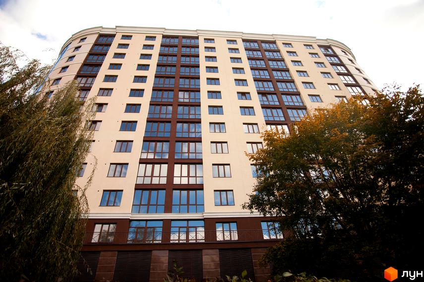 Багатоповерховий житловий комплекс Paradise з вигнутим фасадом у бежевих та коричневих тонах. Навколо будинку видно дерева з зеленим та осіннім листям.