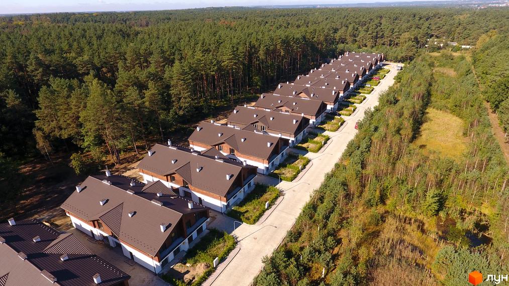 Котеджне містечко КМ Bruhsell: ряд двоповерхових будинків з коричневими дахами та білими фасадами, розташованих серед зеленого лісу. Прибудинкова територія з доглянутими газонами та бруківкою.