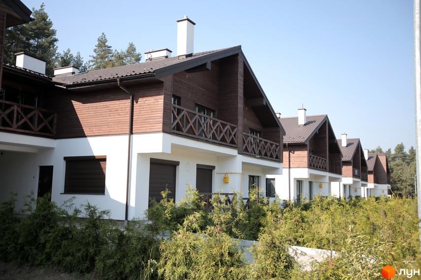 Ряд котеджів Котеджного містечка Bruhsell з білими та коричневими фасадами, балконами з дерев'яними поручнями. На передньому плані зелені кущі, на задньому - сосни.