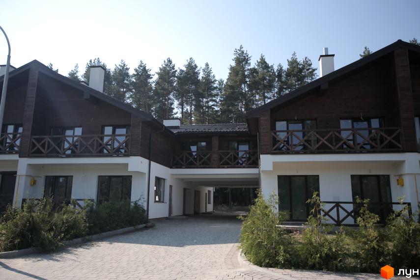 Котеджне містечко Bruhsell: двоповерхові будинки з темними дерев'яними фасадами та балконами, оточені соснами. Прибудинкова територія з бруківкою та зеленими насадженнями.