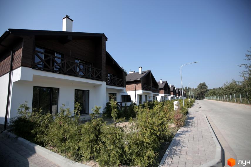 Котеджне містечко Bruhsell: ряд двоповерхових будинків з білими фасадами, темними дахами та балконами. Перед будинками зелені насадження, дорога та тротуар.