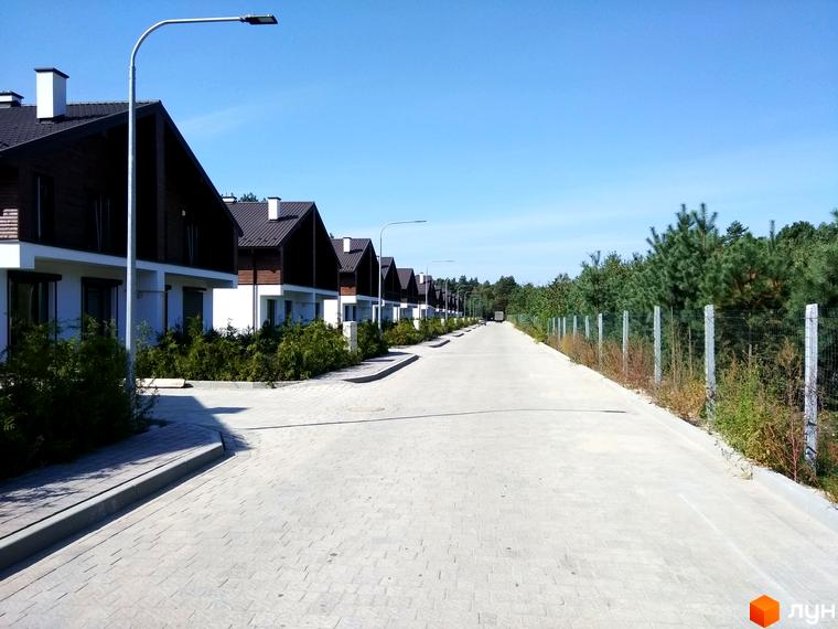 Ряд двоповерхових будинків з темними дахами та світлими фасадами в Котеджному містечку Bruhsell. Перед будинками прокладена дорога та зелені насадження, поруч ліс.