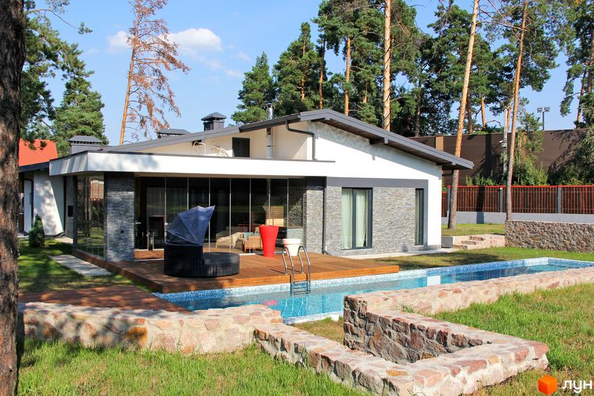 Одноповерховий будинок з великими вікнами та терасою з дерев'яним настилом. На території є басейн та доглянутий газон.