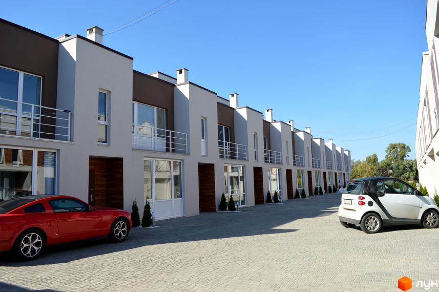 Таунхауси Belvedere City з білими балконами та коричневими вставками на фасаді. Перед будинками розташована брукована територія з червоним та білим автомобілями.