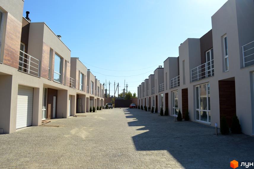 Зображення таунхаусів «Бельведер Сіті». Вигляд на ряд будинків з гаражами та невеликими балконами, розташованих вздовж вимощеної бруківкою дороги.