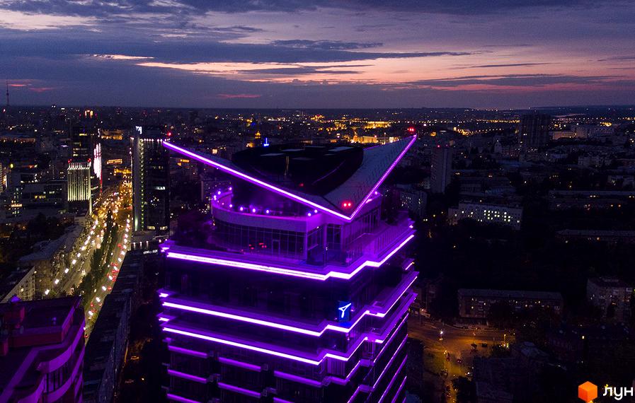 Житловий комплекс Jack House у Києві з яскравою фіолетовою підсвіткою вночі. Вид згори на висотну будівлю з унікальною архітектурою та міським пейзажем на заході сонця.