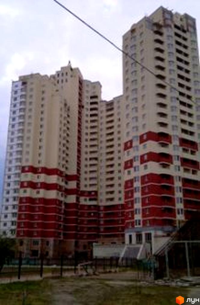 Житловий комплекс Княжий Затон, 9, має світлі фасади з червоними горизонтальними смугами та балконами. На передньому плані видно ділянку з ґрунтом та металевими конструкціями.