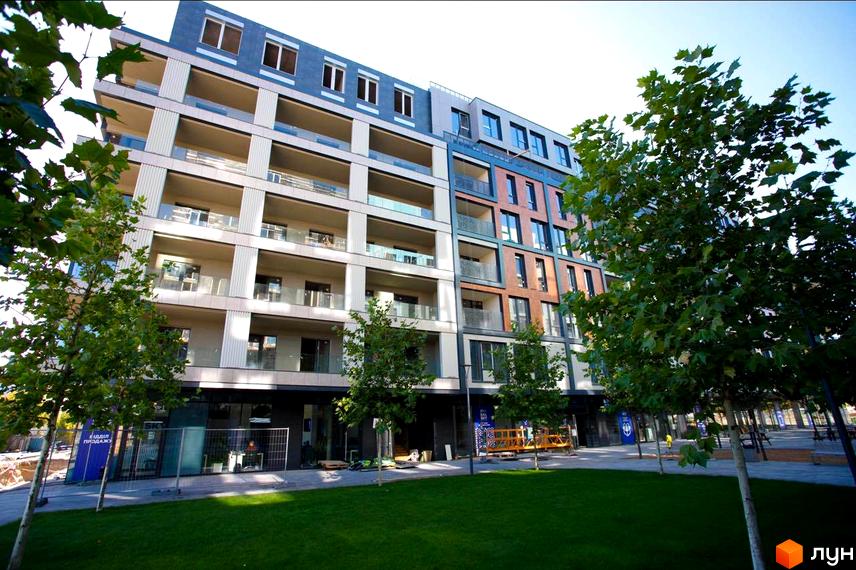 Фасад багатоповерхового будинку Житловий район Rybalsky з світлими та цегляними елементами, скляними балконами. Перед ним зелений газон та молоді дерева.