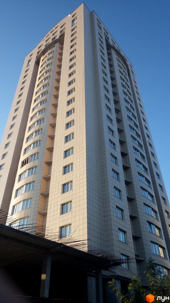 Високий житловий будинок світлого відтінку з багатьма вікнами та вигнутими секціями фасаду. Знизу видно будівельні конструкції.