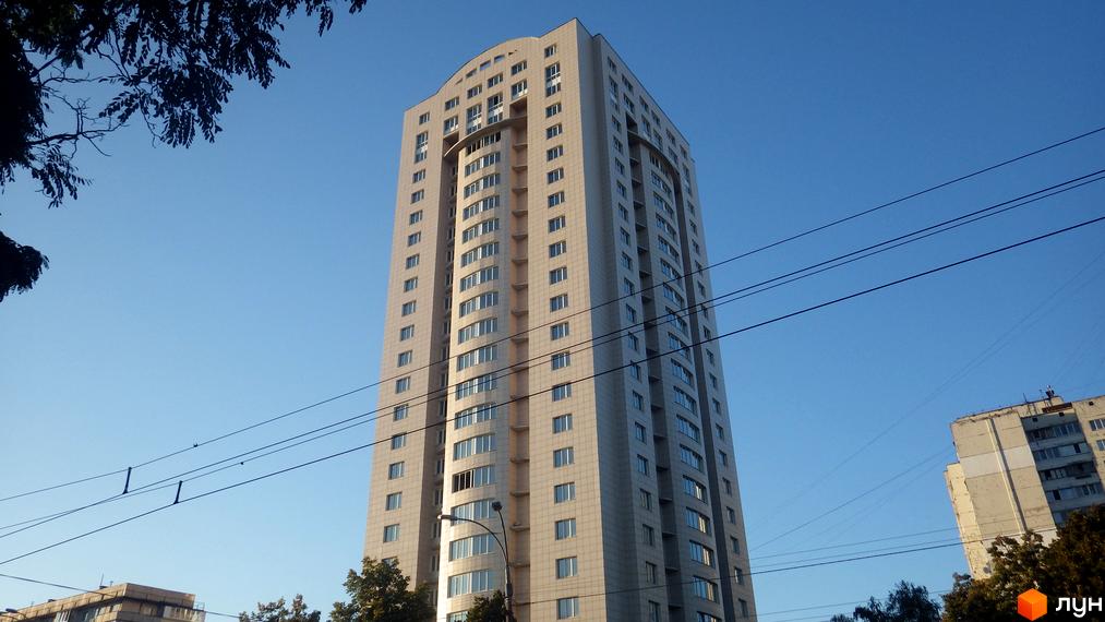 Багатоповерховий житловий комплекс світлого кольору з численними вікнами та вигнутим дахом. На передньому плані видно електропроводи та дерева.