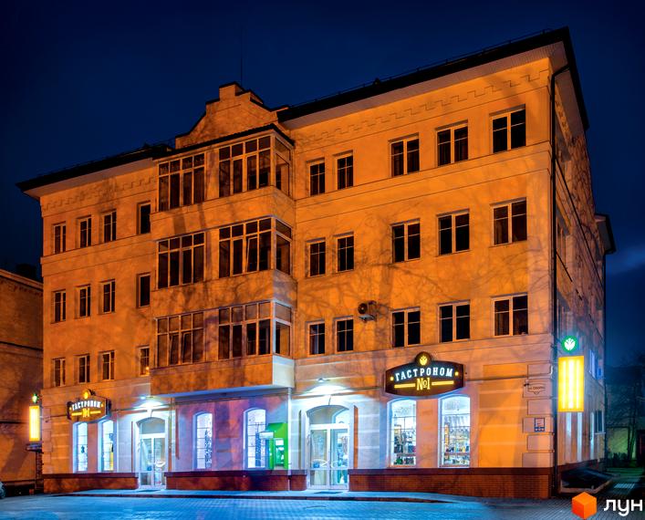 Зовнішній вигляд триповерхового будинку на вулиці Соборна, 217, з освітленим фасадом та вітринами магазинів на першому поверсі. Навколо будинку облаштований тротуар.