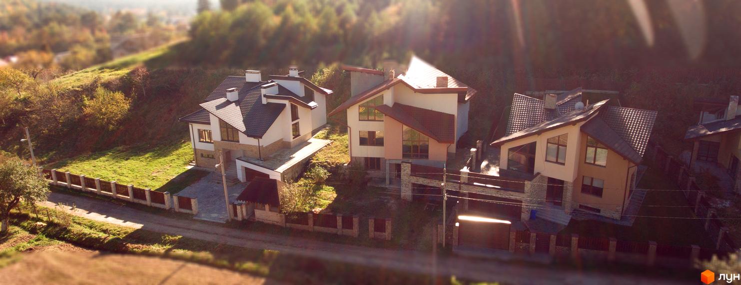 Котеджне містечко КМ Буковий Гай. Сучасні будинки з коричневими дахами та великими вікнами розташовані на схилі пагорба серед дерев. Навколо зелена трава та дерев'яний паркан.