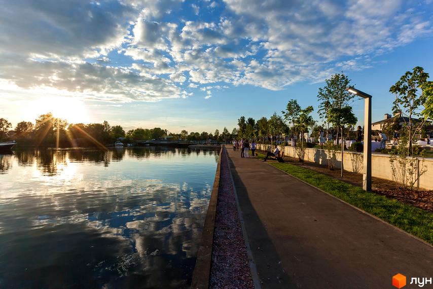 На фото зображено берег водойми з пішохідною доріжкою та ліхтарями. На задньому плані видно будівлі та зелені насадження. КМ Золоче - ідеальне місце для життя.