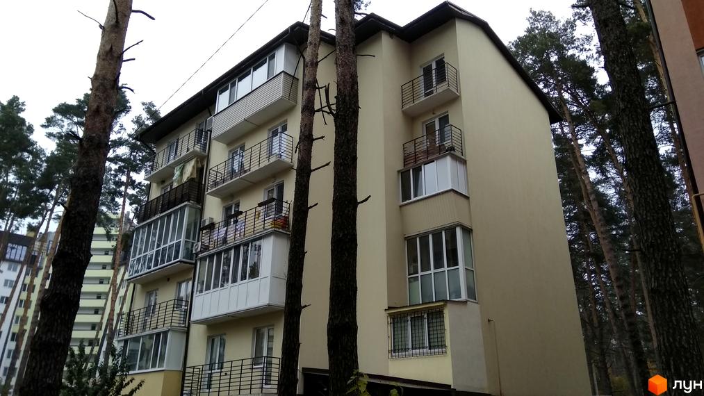 Зовнішній вигляд клубного будинку „10-а лінія“ зі світло-жовтим фасадом та численними балконами. Навколо будівлі ростуть високі сосни.