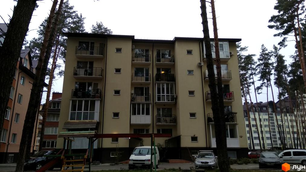 Клубний будинок „10-а лінія“ з балконами та вікнами на тлі дерев. Перед будинком розташована дитяча гірка та припарковані автомобілі.