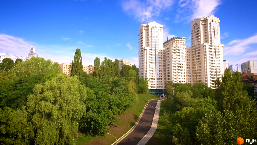 Високі будинки ЖК Dynasty на тлі блакитного неба. Доглянута прибудинкова територія з зеленими насадженнями та дорогою.