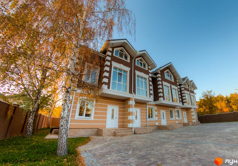 Зображення таунхаусів „Лісовий Маєток“ з коричневими дахами та великими вікнами. На передньому плані дерева з жовтим листям та вимощена плиткою територія.