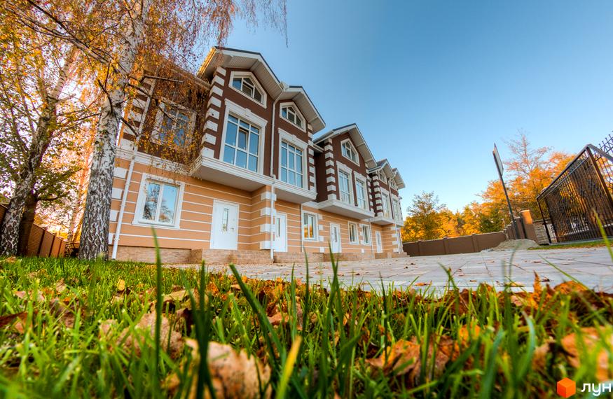 Зображення таунхаусів „Лісовий Маєток“ з фасадом у бежево-коричневих тонах. Перед будинками зелений газон з опалим листям та вимощена доріжка.