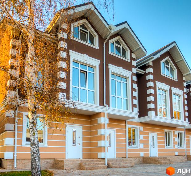 Зображення таунхаусів „Лісовий Маєток“ з коричневими фасадами та великими вікнами. Перед будинками росте дерево з жовтим листям.