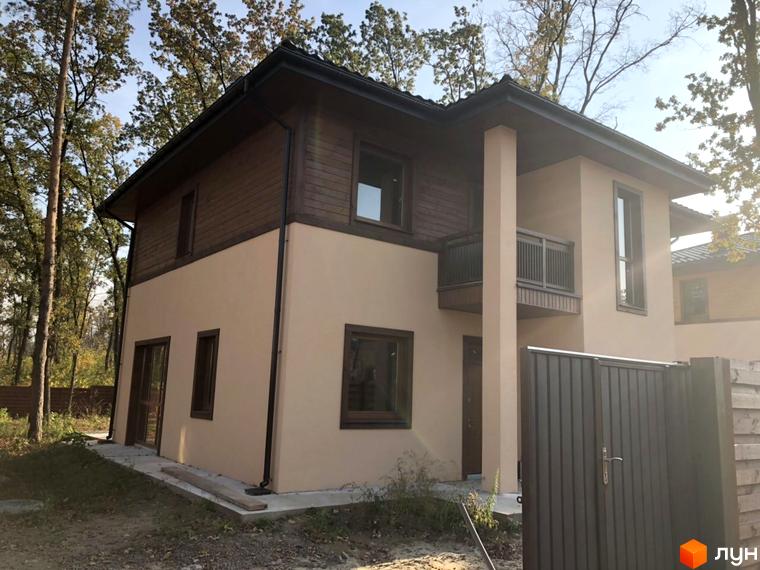 Котеджне містечко Віта Паркова: двоповерховий будинок з бежевим фасадом та дерев'яними елементами, огороджений металевим парканом. Навколо будинку дерева та зелень.