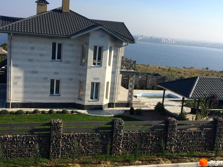 Двоповерховий будинок з сірим дахом та світлим фасадом. На території є газон, басейн та альтанка, а також паркан з габіонами.