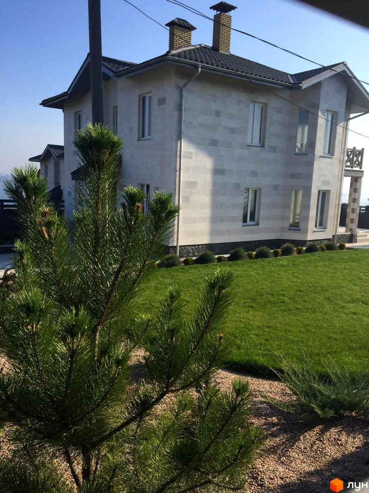 Зовнішній вигляд будинку з двосхилим дахом. На прибудинковій території газон з зеленої трави, декоративні кущі та хвойні дерева.