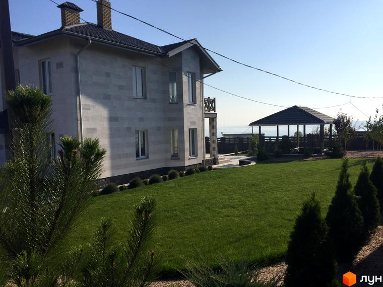 Двоповерховий будинок з світлим фасадом та газоном. На території розташована альтанка та зелені насадження.