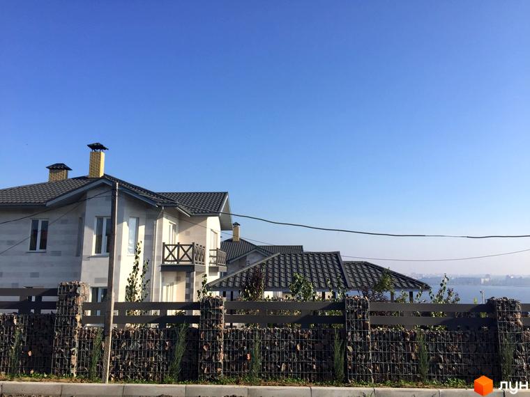 Зображення житлового будинку з сірим дахом та світлими стінами. Перед будинком розташований паркан з каменю та металу.