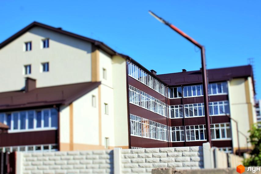 Зовнішній вигляд будинку житлового комплексу Belobrovski з коричневим дахом та великою кількістю вікон. На передньому плані видно кам'яний паркан.