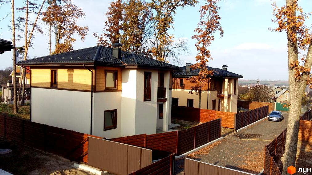 Котеджне містечко КМ Віта Паркова. Двоповерхові будинки з дерев'яними та білими фасадами, чорними дахами, огороджені дерев'яним парканом. Біля будинків розташована парковка та дерева з осіннім листям.