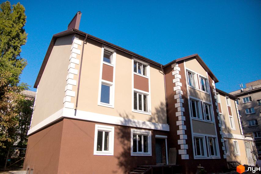 Житловий комплекс Дім на Набережній. Фасад будинку оздоблений бежевими та коричневими елементами з білими декоративними вставками. Навколо будинку зелені насадження.