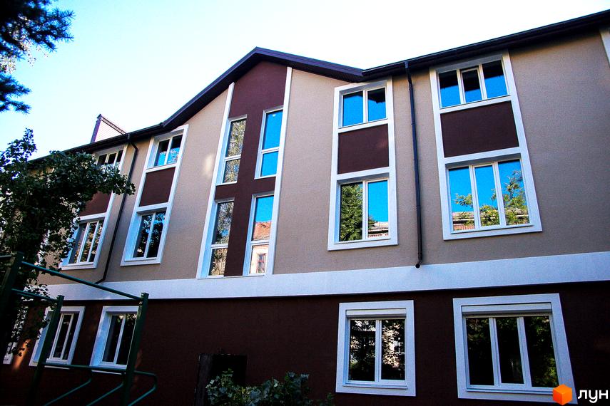 Житловий комплекс Дім на Набережній. Фасад будівлі з коричневими та бежевими елементами, великими вікнами з блакитними склопакетами. Навколо зелені дерева та частина прибудинкової території.