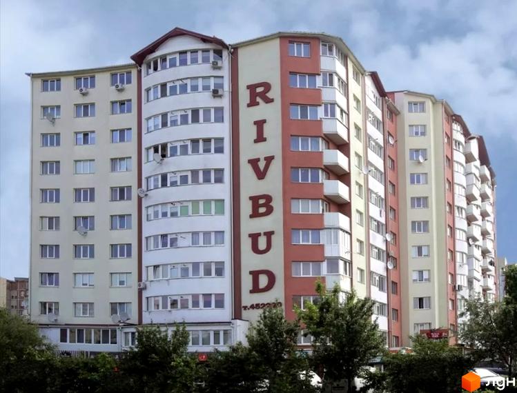 Житловий комплекс Rivbud: багатоповерховий будинок з кремовими та теракотовими фасадами, вигнутою центральною частиною та зеленими деревами.