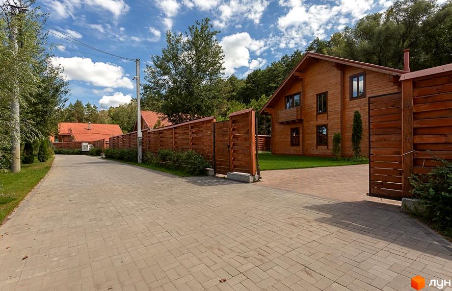 Котеджне містечко Summerville: двоповерховий дерев'яний будинок з червоним дахом та дерев'яною огорожею. Територія перед будинком вимощена плиткою, оточена зеленню.