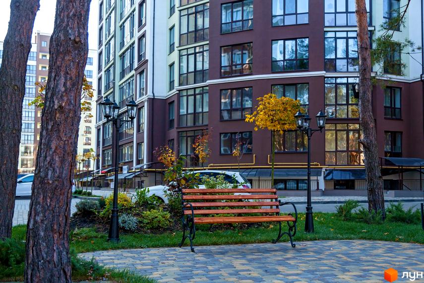 Зображення фасаду та прибудинкової території Житлового комплексу Лісова казка. На фото видно лавочку, дерева та ліхтарі.