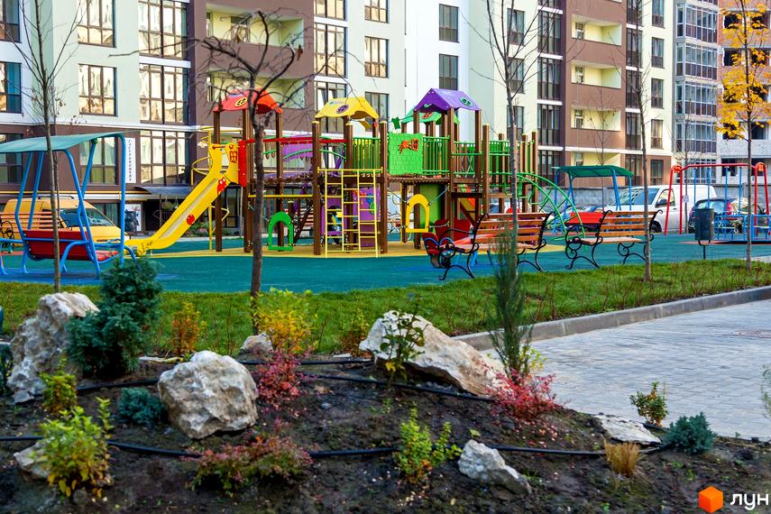 Зображення дитячого майданчика біля багатоповерхового будинку в Житловому комплексі Лісова казка. На території є гірки, гойдалки та лавки.