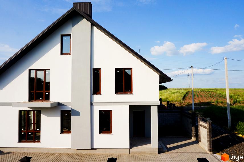 Білосніжний будинок з коричневими вікнами на тлі блакитного неба. На території видно бруківку та зелену рослинність.