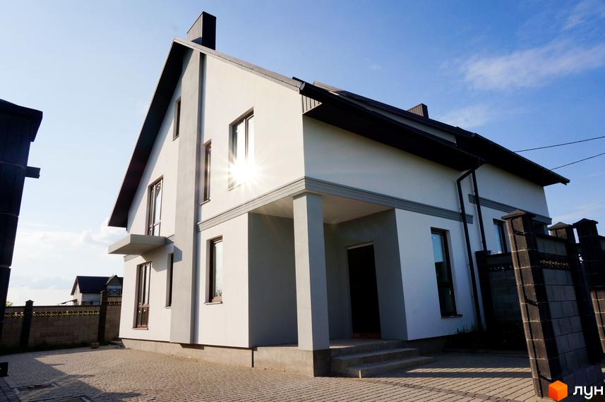 Двоповерховий дуплекс Дуплекси „Білий корал“ з білими стінами та темним дахом. Перед будинком бруківка та бетонна огорожа.