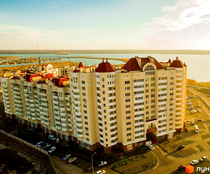 Житловий комплекс Рів'єра з червоними дахами та світлими фасадами на тлі річки. Навколо комплексу розташовані дороги та паркування з автомобілями.