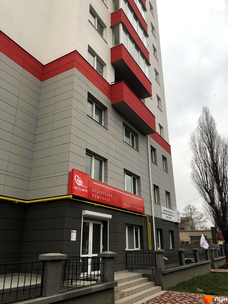 Фасад новобудови ЖК Плаза Квартал з червоними та сірими елементами. Поруч розташована прибудинкова територія із зеленими насадженнями та огорожею.