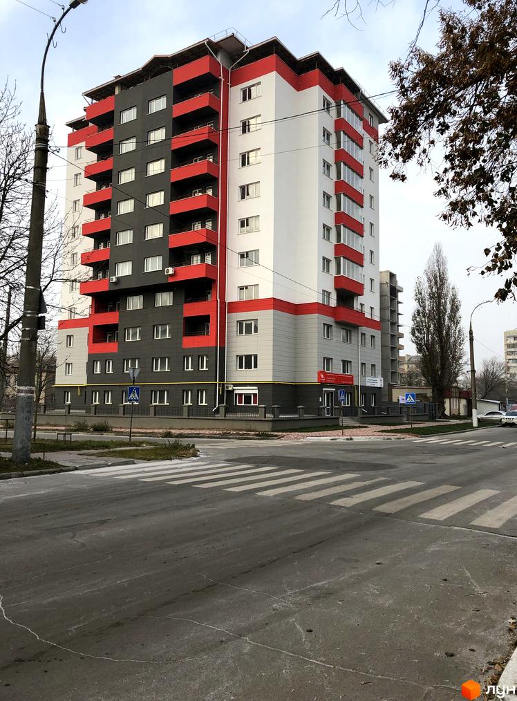 Житловий комплекс Плаза Квартал – висотна будівля з білими, сірими та червоними фасадами та балконами. Поруч дорога з пішохідним переходом.